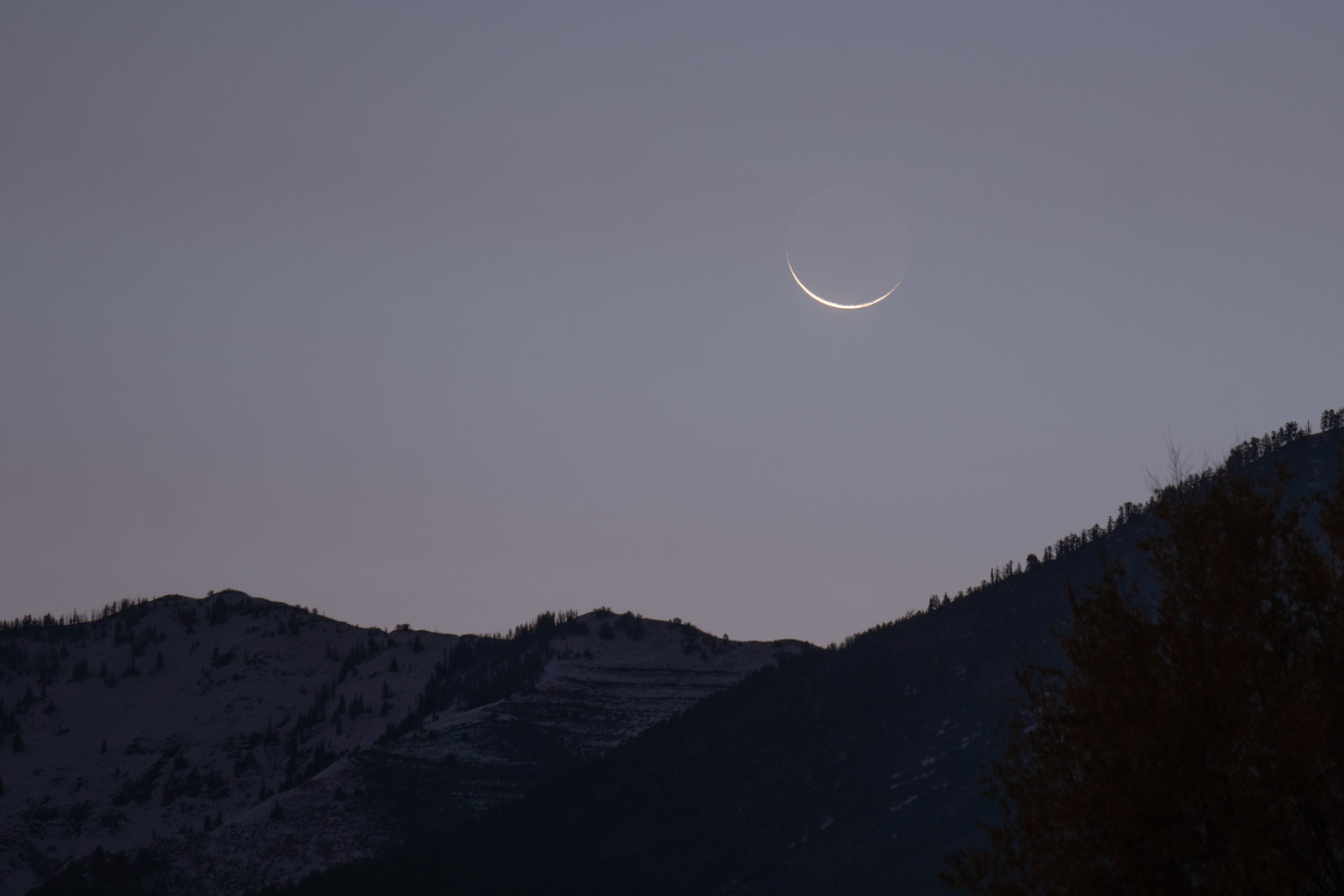 The barely visible thumb nail crescent just about to fall behind a snowy mountain